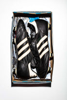 El par de botas sin acordonar en su caja original que conserva la llave de recambio de tacos y la bolsa adidas en su interior.